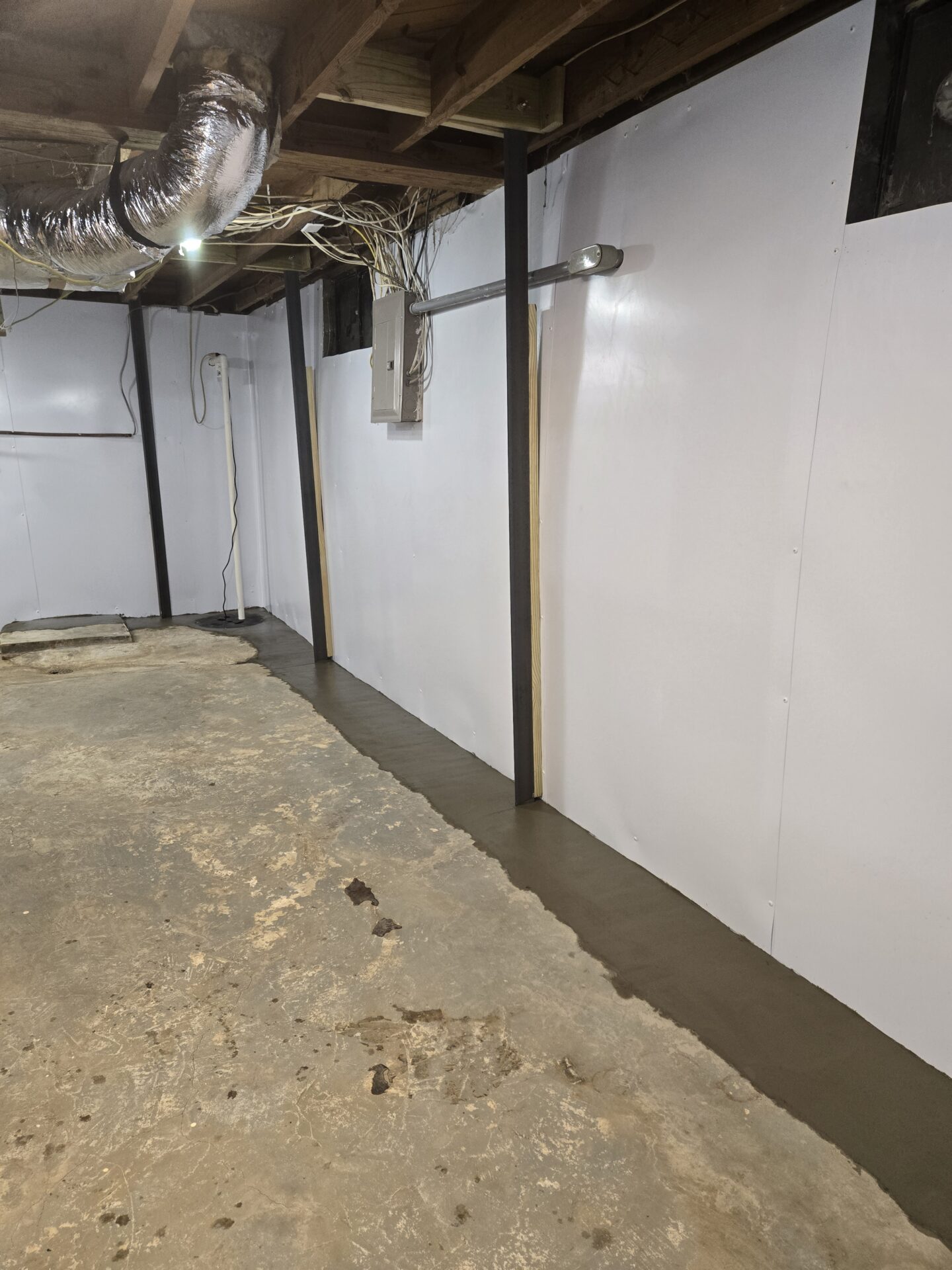 Basement corner with exposed ductwork, beams, and wiring against a white ceiling and concrete wall